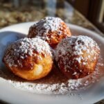 Three golden brown, fluffy apple fritters generously dusted with powdered sugar on a white plate.