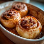 Close-up of three golden brown Baked Cinnamon Apples, glistening with syrup and filled with a spiced apple mixture, in a white baking dish.