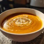 A close-up of a white bowl filled with vibrant orange butternut squash soup, topped with a swirl of cream and black pepper.