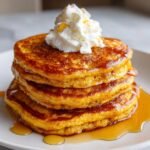 A stack of fluffy pumpkin pancakes topped with whipped cream and a drizzle of syrup.