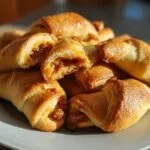 A close-up of a pile of golden-brown baked rugelach on a white plate, with one rugelach broken open to show the filling.