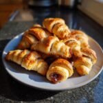 A pile of freshly baked, golden brown rugelach with a sweet filling, presented on a light gray plate.