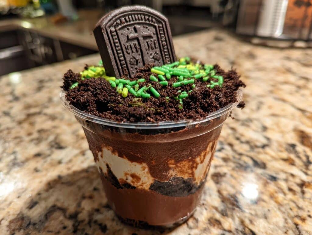 A Halloween Graveyard Pudding Cup topped with crushed cookies, green sprinkles, and a chocolate tombstone cookie.