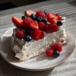 A slice of Holiday Pavlova with Berries, featuring crisp meringue, whipped cream, strawberries, raspberries, and blueberries.