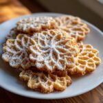 A stack of golden brown Italian Pizzelle Cookies, dusted with powdered sugar, on a white plate.
