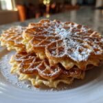 A stack of golden-brown Italian Pizzelle Cookies dusted with powdered sugar on a white plate.