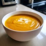 A bowl of creamy orange soup, likely a sweet potato or squash soup, swirled with cream. This is a Maple Pecan Pie inspired soup.