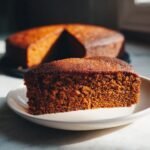 A slice of moist Gingerbread Cake on a plate, with the rest of the cake in the background.