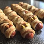 Close-up of several Mummy Hot Dogs wrapped in pastry with candy eyes, ready for a spooky party.