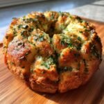 A golden-brown Savory Herb Monkey Bread, generously topped with fresh green herbs, on a wooden cutting board.