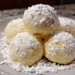 A close-up of a stack of perfect snowball cookies generously dusted with powdered sugar on a light gray plate.