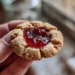 A close-up of a thumbprint jam cookie held in a hand, filled with glistening raspberry jam.