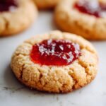 Close-up of a delicious Thumbprint Jam Cookie with glistening red jam in the center and a sugar-dusted cookie base.