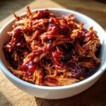 Close-up of shredded bbq chicken crockpot in a white bowl, glistening with sauce.