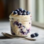 Close-up of Blueberry Cheesecake Overnight Oats in a glass jar, topped with fresh blueberries and a spoon.