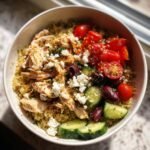 A vibrant Greek Chicken Quinoa Bowl topped with shredded chicken, cherry tomatoes, cucumber slices, Kalamata olives, and feta cheese.