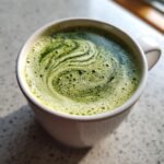 Close-up of a frothy lavender matcha drink in a white mug with beautiful green swirls.
