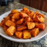 A close-up of a white plate filled with perfectly roasted sweet potato cubes, seasoned and slightly caramelized.