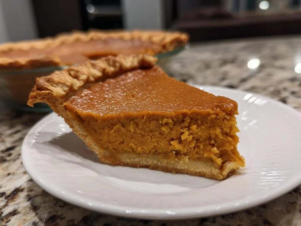 A close-up of a perfect slice of sweet potato pie on a white plate, with the rest of the pie in the background.