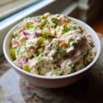 A close-up of a creamy tuna salad recipe with chopped celery, red onion, and chives in a white bowl.