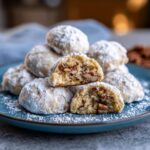 A stack of Buttery Pecan Snowball Cookies on a blue plate, one cookie is halved to show the pecan filling.