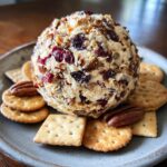 A delicious Cranberry Pecan Cheese Ball on a plate with crackers and pecans.