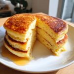 A stack of fluffy lemon ricotta pancakes with a slice cut out, drizzled with syrup on a white plate.