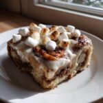 A single hot cocoa cinnamon roll topped with icing and toasted marshmallows on a white plate.