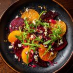 Overhead shot of a vibrant Beet and Orange Salad with arugula and crumbled cheese on a dark plate.
