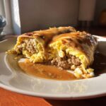 Two Breakfast Enchiladas with Sausage Gravy on a plate, filled with sausage and eggs and covered in gravy.