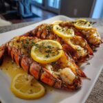 Three butter poached lobster tails on a white plate, garnished with lemon slices and thyme.