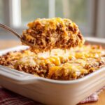 Spoon lifting a portion of cheesy ground beef rice casserole from a baking dish, showing melted cheese and layers.