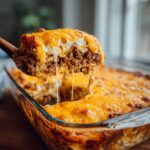 A wooden spoon lifts a serving of Crazy Good Casserole from a glass baking dish, showcasing a cheesy, layered texture.