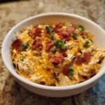 A bowl of Crock Pot Crack Chicken topped with melted cheese, bacon bits, and green onions.