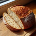 A loaf of crusty German Bread with a slice cut, sitting on a wooden board in natural light.