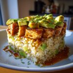 A delicious Honey Lime Chicken Avocado Rice Stack on a white plate, drizzled with sauce and garnished with herbs.
