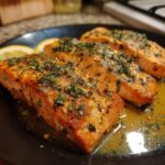Three fillets of Lemon Garlic Salmon on a black plate, garnished with herbs and lemon slices.