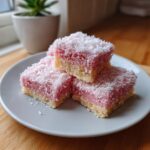 Three Pink Coconut Snowball Cake Bars stacked on a white plate, topped with shredded coconut.