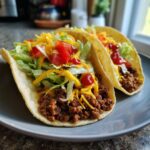 Two smash burger tacos filled with seasoned meat, lettuce, tomatoes, cheese, and sauce on a gray plate.