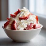 A bowl of Tres Leches Whipped Cream Strawberries, topped with fresh whipped cream and sliced strawberries.