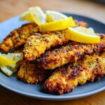 A plate of Zesty Lemon Pecorino Crispy Chicken garnished with lemon wedges. Crispy, golden-brown chicken tenders.