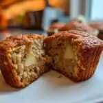 Close-up of an Apple Cinnamon Muffin cut in half, showing the apple pieces inside. Other muffins are in the background.