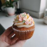 A delicious baby in bloom cupcake held in hand, decorated with frosting and small pink, yellow, and green flower sprinkles.