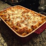 A red casserole dish filled with baked mostaccioli, topped with melted cheese and herbs.