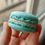 Close-up of a blue Macarons held between two fingers, showing the filling and textured shell.