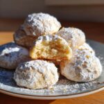 A stack of Condensed Milk Snowball Cookies dusted with powdered sugar on a blue plate, one cookie broken in half.