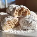 A pile of Condensed Milk Snowball Cookies, one broken in half, dusted with powdered sugar on a white plate.