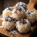 A pile of homemade Condensed Milk Snowballs covered in coconut flakes and drizzled with chocolate on a wooden board.