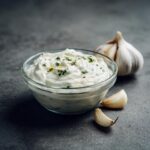Close-up of creamy Garlic Aioli in a glass bowl, garnished with herbs, next to garlic cloves.