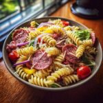 A bowl of grinder pasta salad with salami, tomatoes, red onion, cheese, and spiral pasta.